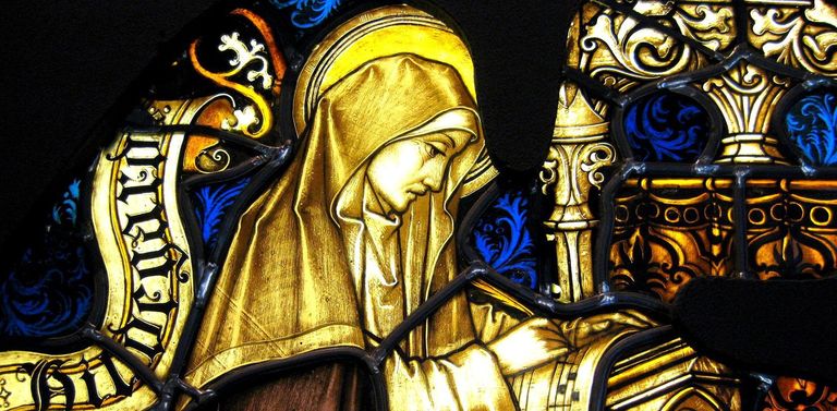 Hildegard von Bingen im Ordensgewand und mit Heiligenschein als Teil eines Kirchenfensters. Sie beschreibt ein Blatt Notenpapier mit einer Feder.